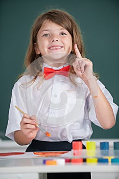 Child girl draws in classroom sitting at a table, having fun on school blackboard background. Portrait of little girl