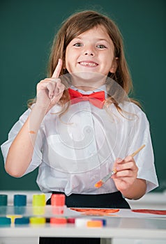Child girl draws in classroom sitting at a table, having fun on school blackboard background. Portrait of little girl