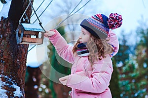 child girl feeding birds in winter. Bird feeder in snowy garden, helping birds during cold season