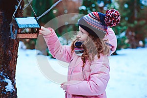 Child girl feeding birds in winter. Bird feeder in snowy garden, helping birds during cold season