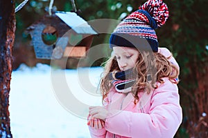 Child girl feeding birds in winter. Bird feeder in snowy garden, helping birds during cold season