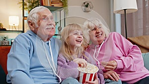 Child girl granddaughter grandfather grandmother eating popcorn watching movie film on home sofa