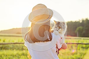 Child girl in hat with gray fluffy cat in her arms. Beautiful sunset country landscape background