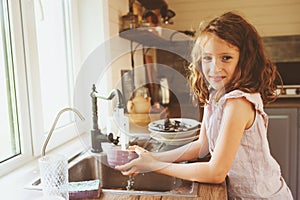 Child girl helps mother at home and wash dishes in kitchen. Casual lifestyle in real interior