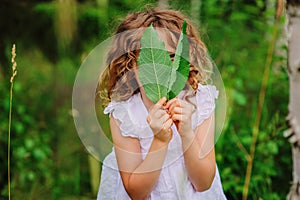 Child girl hiding behind green leaves in summer forest