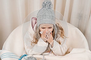 Child girl at home in chair with warm knitted hat woolen blanket sneezes in handkerchief. Season autumn winter colds photo