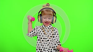 Child girl listening music via headphones, working out pumping up arm muscles lifting pink dumbbells