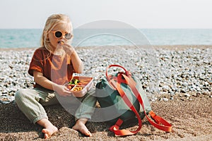 Child girl with lunchbox eating vegetables outdoor travel vacation healthy lifestyle vegan food picnic on beach hungry kid
