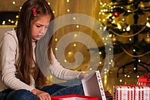 Child girl opening Christmas gift in decorated room and gets upset