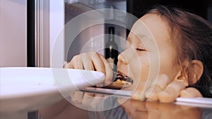 Child girl is opening a fridge, taking an eclair and eating it furtively.