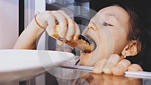 Child girl is opening a fridge, taking an eclair and eating it furtively.