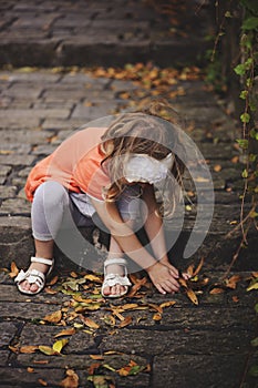 Child girl in orange cardigan gathering leaves on old stone stairs