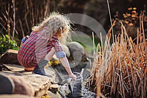 Child girl in plaid dress gathering water from pond in spring garden