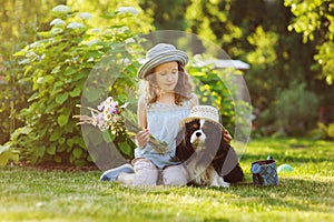 child girl playing with her spaniel dog in summer garden, both wearing funny gardener hats
