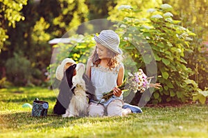Child girl playing with her spaniel dog in summer garden, both wearing funny gardener hats, holding bouquet