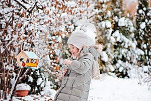 Child girl puts seeds in bird feeder in winter snowy garden