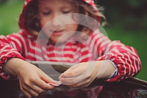 Child girl in red raincoat playing with water barrel, rainy day outdoor activities