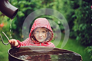 Child girl in red raincoat playing with water barrel in rainy summer garden. Water economy and nature care