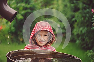 Child girl in red raincoat playing with water barrel in rainy summer garden. Water economy and nature care