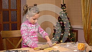 Child girl rolls out cookie dough for Christmas in the kitchen. The child helps his mother to cook on the background of