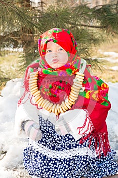 Child girl in Russian pavloposadskie folk scarf on head with floral print and with bunch of bagels on background of snow.