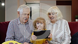 Child girl shows something in laptop to grandparents, seniors couple learning how to use tablet