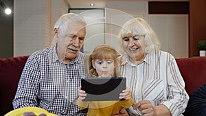 Child girl shows something in laptop to grandparents, seniors couple learning how to use tablet