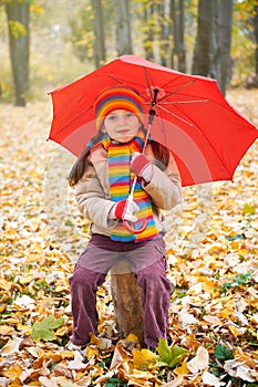 Child girl sit on trunk with red umbrella in autumn forest, fallen leaves on background, fall season