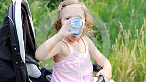 Child girl sits in a baby stroller and drinks water from a bottle.