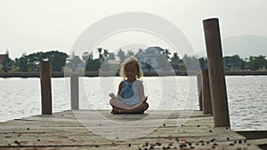 Child girl sits on the wood pier in lotus pose and meditate in slow motion