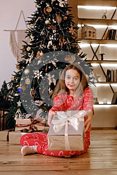 Child girl sitting near a Christmas tree in red pajamas and holding a Christmas present