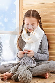 Child girl is sitting on a window sill and knitting the scarf from gray wool yarn. Beautiful view outside the window - sunny day
