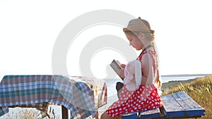 Child girl in a straw hat and dress sitting on bench and reading book. Cute kid with soft rabbit toy looking at notebook