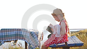 Child girl in a straw hat and dress sitting on bench and reading book. Cute kid with soft rabbit toy looking at notebook