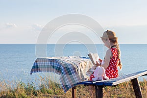 Child girl in a straw hat and dress sitting on bench and reading book. Cute kid with soft rabbit toy looking at notebook