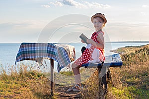 Child girl in a straw hat and dress sitting on bench and reading book. Cute kid with soft rabbit toy looking at notebook