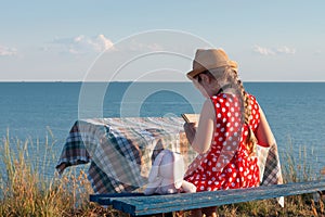 Child girl in a straw hat and dress sitting on bench and reading book. Cute kid with soft rabbit toy looking at notebook