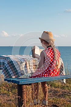 Child girl in a straw hat and dress sitting on bench and reading book. Cute kid with soft rabbit toy looking at notebook