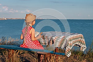 Child girl in a straw hat and dress sitting on bench and reading book. Cute kid with soft rabbit toy looking at notebook