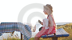 Child girl in a straw hat and dress sitting on vintage bench and reading a book. Cute kid with soft rabbit toy looking at notebook
