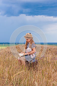 Child girl in a straw hat and dress sitting on vintage suitcase and reading a book.Cute kid with toy looking at notebook