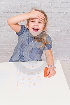 Child, a girl at the table writes, draws on a piece of paper, against a white brick wall