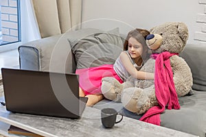 Child girl watching laptop screen, hugging teddy bear. Concept of distance learning and leisure of modern children at computer