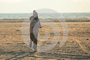 A child girl wearing the hat with a pompon strolling on the beach at sunset