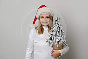 Child girl wearing Santa hat and white t-shirt holds Christmas tree. Xmas kid 10 years old portrait