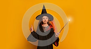 Child girl in wizard costume with luminous magic wand looking at camera and pointing finger up on yellow background.