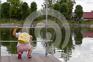 A child girl with a yellow backpack in summer looks at the water in the pond and tries to see the fish there. Leaning