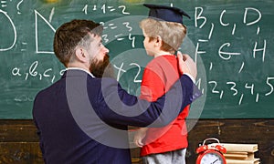 Child in graduate cap listening teacher, chalkboard on background, rear view. Teacher with beard, father teaches little son in