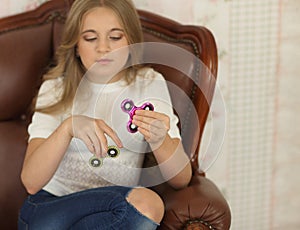Child with green and pink fidget spinners in hands, sitting in leather chair, wearing white shirt and jeans.