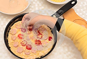 Child hand placing the cheese onto the raw dough for homemade pizza with sausages and tomatoes. Other ingredients are on